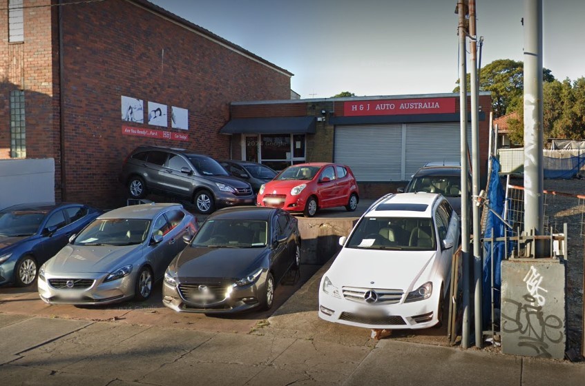 The H & J AUTO AUSTRALIA sign over a roller door at 60 Parramatta Road, Croydon, with cars parked nose-in along the footpath in front of the brick building