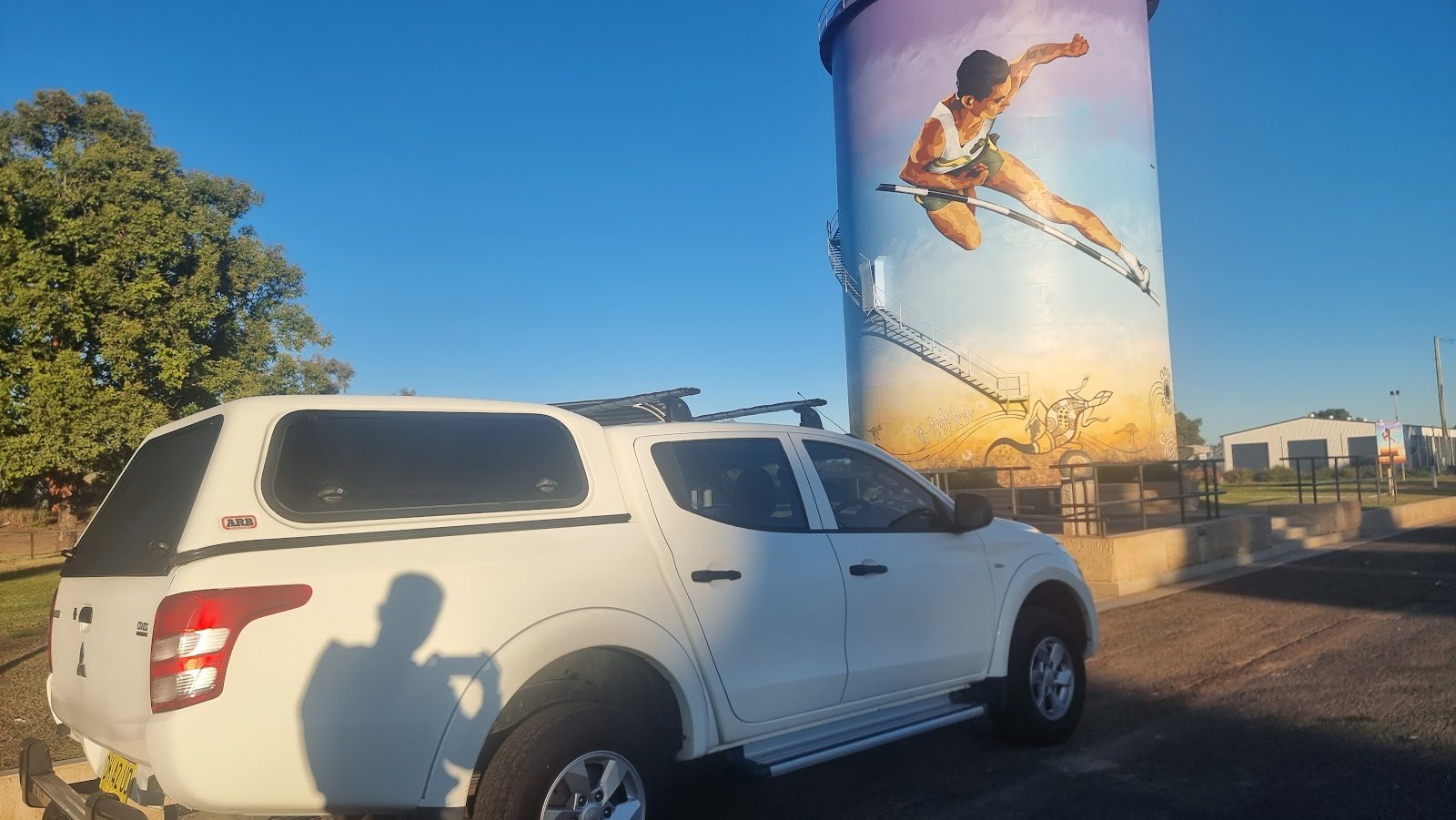The same white Triton in late sun beside a water tower painted with a hurdler